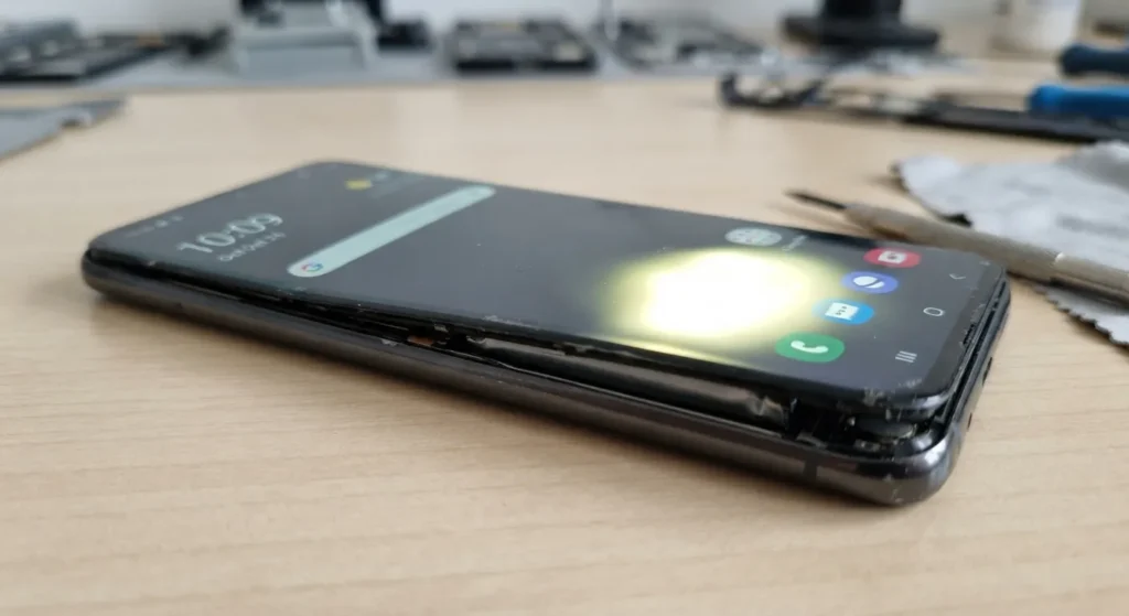 Damaged smartphone with a lifted screen exposing a swollen battery on a repair desk.